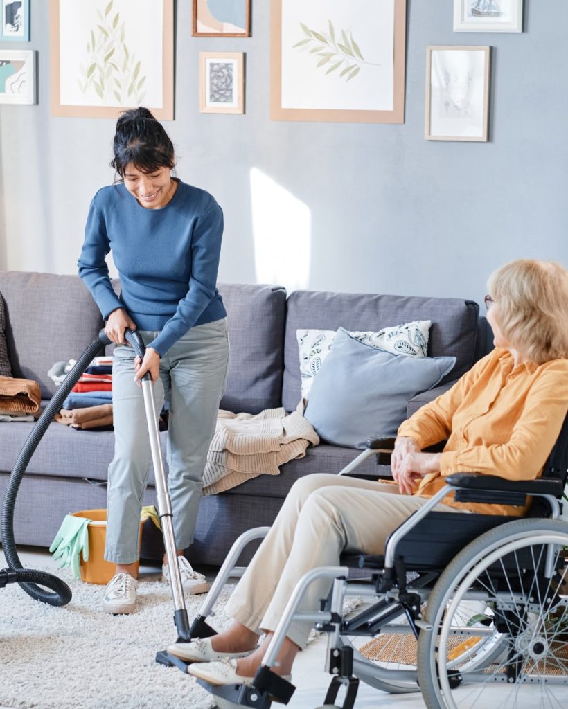 senior-woman-using-wheelchair-talking-housekeeper-who-vacuuming-floor-room (Large)